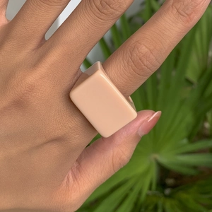 Détail d'une bague carrée large en résine orange pêche portée sur une main avec plantes