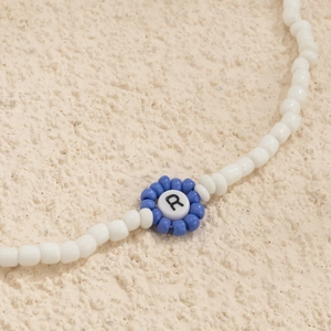 Vista macro de collar de cuentas blancas con flor azul y detalle de la letra R central