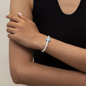 Primer plano de una pulsera de cuentas blancas con flor azul y letra H impresa al centro