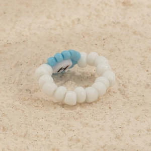 Detalle de anillo de perlas hecho a mano con flor azul y letra H sobre fondo de arena