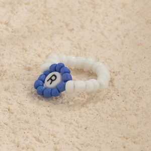 Detalle de un anillo artesanal de cuentas azules y blancas con un medallón de la letra R