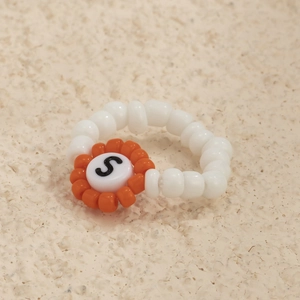 Detalle de anillo de cuentas blancas con flor naranja y la letra S grabada