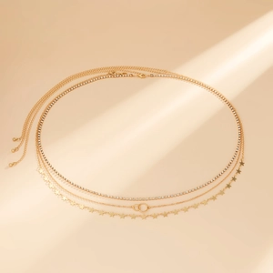 Détail d'une chaîne de corps triple dorée avec strass et breloques étoiles sur fond beige