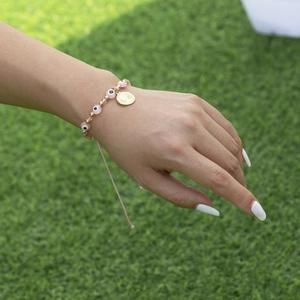 Bracelet tressé œil maléfique rose avec pendentif pièce d'or porté sur herbe verte
