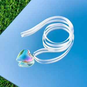 Iridescent heart pendant with white satin ribbon on a mirror background with blue sky reflection