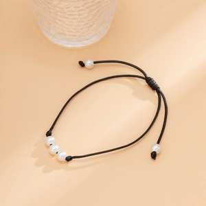 Detalle de pulsera de cordón negro ajustable con tres perlas blancas sobre superficie melocotón