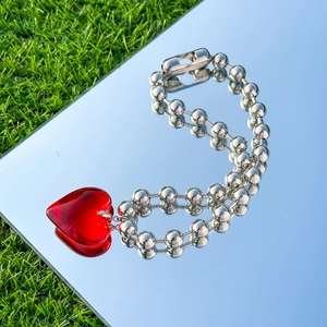 Close-up of red resin heart pendant on silver beaded chain over a mirror and grass