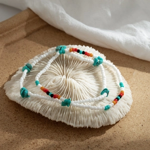 Detalle de tobilleras bohemias con cuentas blancas y toques de turquesa sobre coral blanco
