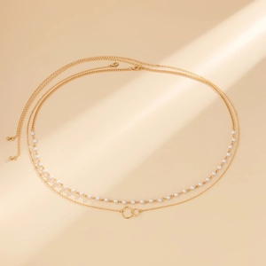Detail einer mehrlagigen goldenen Körperkette mit Perlen und Ringen auf beigem Hintergrund