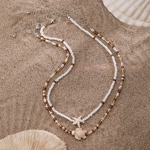 Detail of layered beach necklace with starfish and turtle pendants on sandy background
