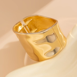 Detail of a wide gold metal bracelet featuring irregular wave textures and a sleek hinge