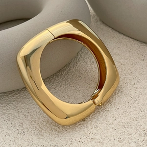 Polished gold square bangle with hinge clasp on a modern stone display base