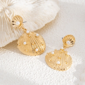 Detail of golden shell drop earrings with white pearls on a textured beach-style background