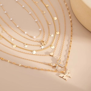 Détail de colliers dorés superposés avec perles, papillons et étoiles sur fond beige clair