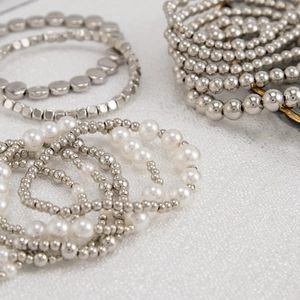 Detail von silbernen Perlenarmbändern mit Herzanhänger und weißen Perlen auf Stoff