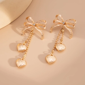 Détail de boucles d'oreilles nœud dorées avec cœurs, gravure florale et chaînes à perles