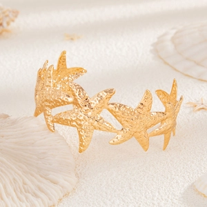 Detailansicht eines goldenen Seestern-Armreifs auf weißem Sand mit echten Muscheln