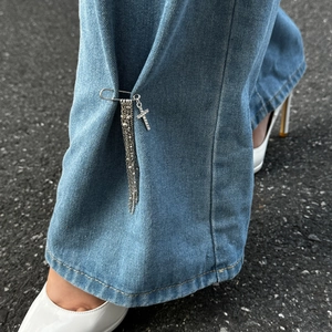 Dettaglio spilla da balia croce argento con nappe su jeans e tacchi a spillo bianchi