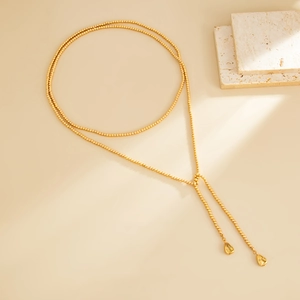 Lange gouden kralenketting in Y-vorm met gepolijste druppelhangers op een beige steen
