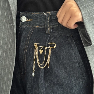 Dettaglio di una spilla punk dorata con catene, ciondolo a cuore e perla su tessuto denim