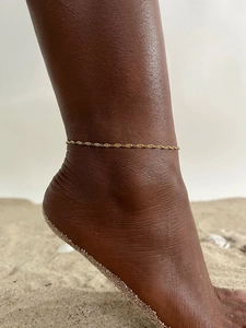 Close-up of a minimalist golden flat snake chain anklet worn on a beach with sand