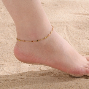 Gouden roestvrijstalen enkelbandje met kleurrijke emaille bedeltjes gedragen op een zandstrand
