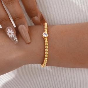 Close-up van een gouden kralenarmband met een fonkelende hartvormige zirkoon op de pols