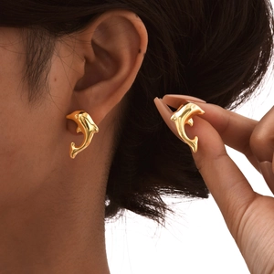 Polished gold dolphin stud earrings for women shown on ear and held by model hand