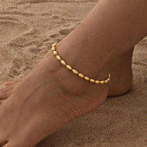 Detail der goldenen Kugelkette am Knöchel einer Frau vor weichem Sandstrand-Hintergrund