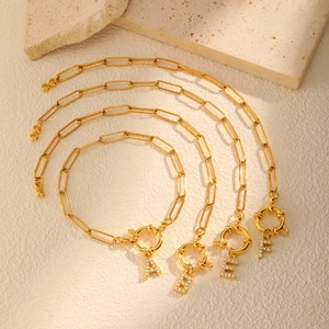 Gouden paperclip ketting met strass letterhangers A, B, E, F op een stenen ondergrond