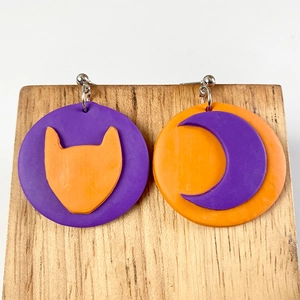 Macro shot of handmade Halloween clay earrings featuring a purple cat and orange moon