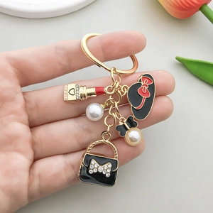 Porte-clés doré avec charms de rouge à lèvres et chapeau noir sur un fond gris épuré