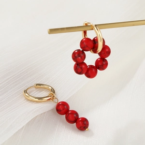 Detail asymmetrischer Goldohrringe mit roten Perlen vor elegantem weißem Stoffhintergrund