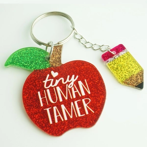 Macro view of glittery apple and pencil keychains with tiny HUMAN TAMER text for teachers