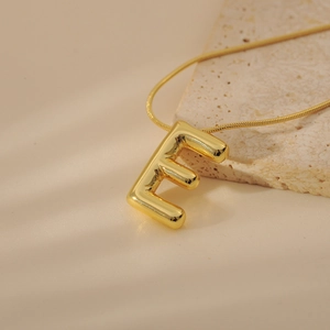 Macro side view of puffy gold letter E pendant on a sleek snake chain over stone