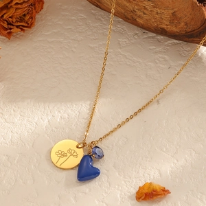 Goldene Edelstahlkette mit Blumenmedaillon und blauem Herzanhänger auf strukturiertem Hintergrund