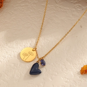 Goldene Halskette mit blauem Herzanhänger und graviertem Blumen-Charm auf weißem Grund