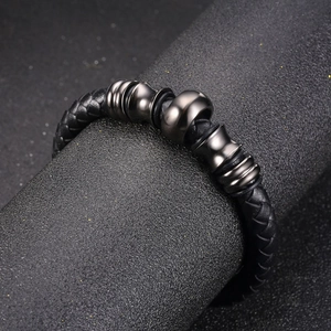 Men's braided black leather bracelet with gunmetal charms on a textured dark pedestal