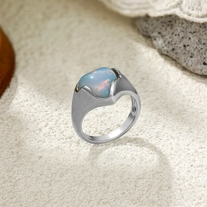 Détail d'une bague mate argentée avec une grande pierre d'opale irisante sur du sable blanc