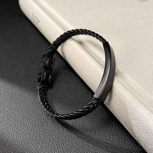Detalle de pulsera de cuero negro trenzado con placa metálica mate y cierre de acero