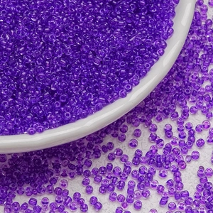 Macro view of translucent 3mm purple glass seed beads scattered near a white porcelain bowl