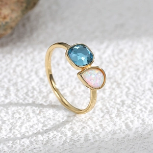 Anillo dorado con piedra azul cuadrada y ópalo de lágrima sobre fondo de piedra gris