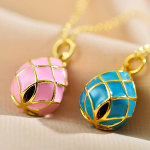 Detailed close-up of pink and blue enamel egg pendants on gold chains over satin