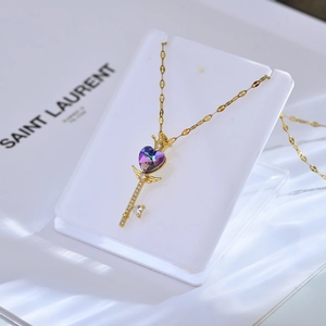 Detail van gouden sleutelketting met paarse hart-zirkonia en kroonvleugels op standaard