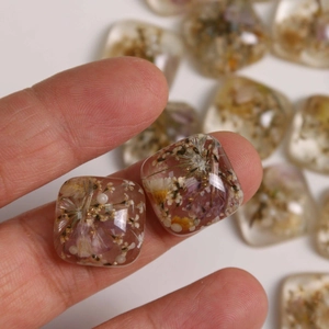 Handheld shot of clear square resin charms with dried flowers and gold foil accents