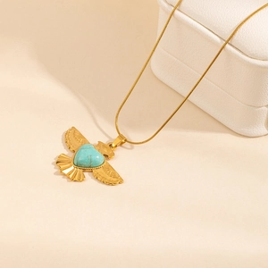 Collana dorata a forma di uccello con cuore di turchese su sfondo beige