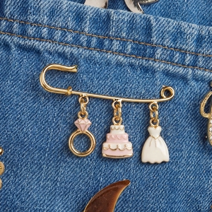 Gold-plated safety pin brooch with wedding ring, cake, and dress charms on denim