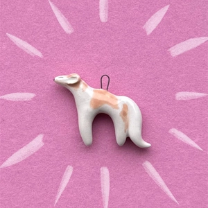 Handmade ceramic greyhound pendant with white and tan spots on a pink felt background