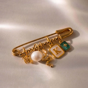 Detalle de un broche de imperdible dorado con perla, colgante de serpiente y letra K