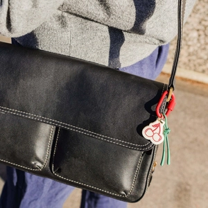 Detail eines Kirsch-Anhängers mit rotem Karabiner und grüner Kordel an einer Ledertasche